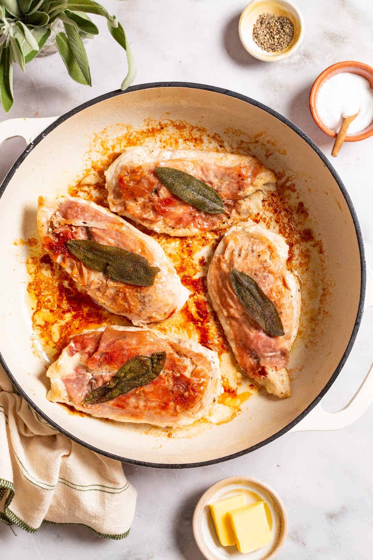 An overhead photo of chicken saltimbocca in a skillet. This is surrounded by sage, a kitchen towel and small bowls of salt, pepper and butter.