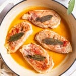 An overhead photo of chicken saltimbocca in a skillet. This is surrounded by sage, a kitchen towel and small bowls of salt, pepper and butter.