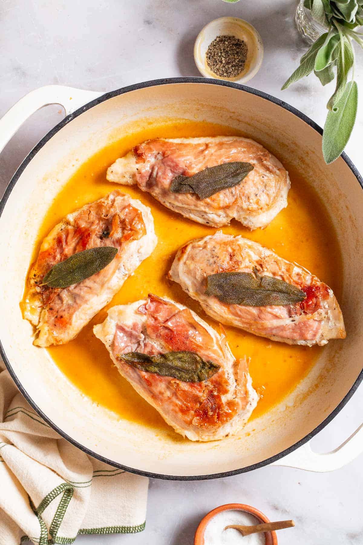 An overhead photo of chicken saltimbocca in a skillet. This is surrounded by sage, a kitchen towel and small bowls of salt, pepper and butter.