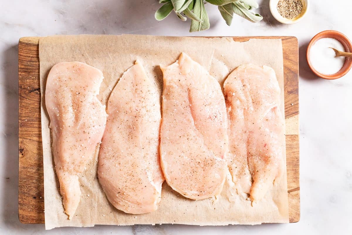 4 uncooked chicken breasts seasoned with salt and pepper on a parchment lined cutting board. Next to this are small bowls of salt and pepper and some sage.