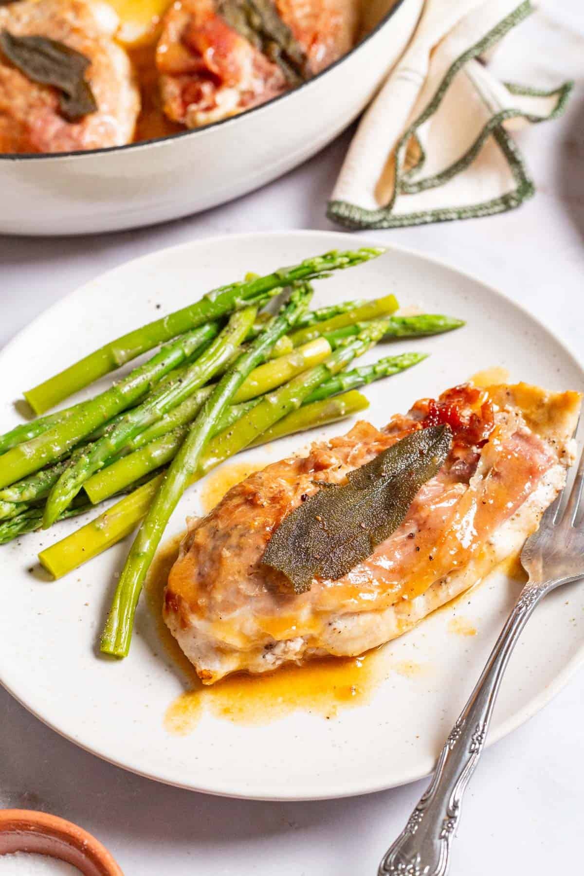 A close up of a serving of the chicken saltimbocca on a plate with a side of asparagus and a fork. Next to this is the rest of the chicken in a skillet and a kitchen towel.