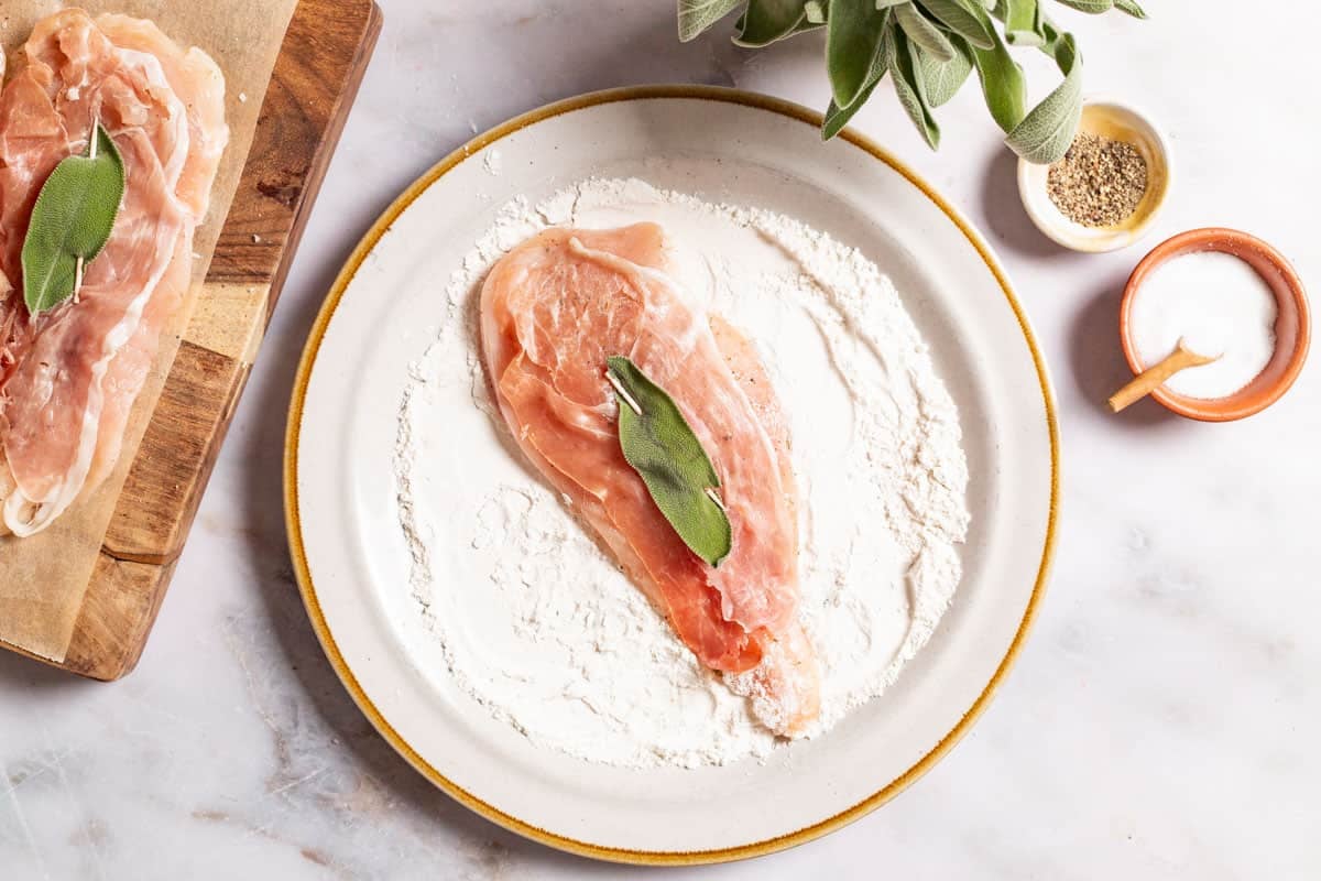 An uncooked chicken breasts seasoned with salt and pepper and topped with a sage leaf being dredged in flour on a plate. Next to this are small bowls of salt and pepper and some sage.