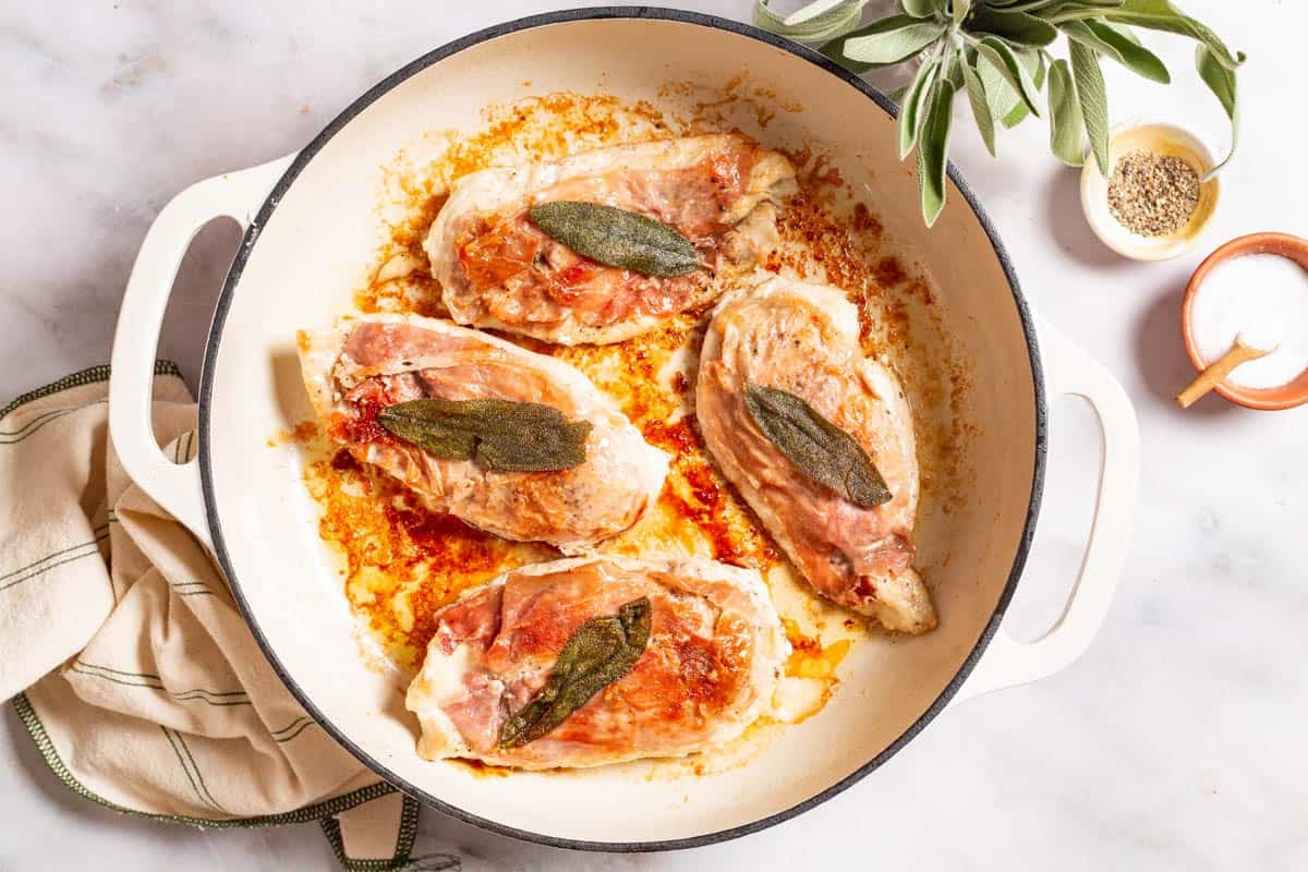 An overhead photo of chicken saltimbocca in a skillet. This is surrounded by sage, a kitchen towel and small bowls of salt, pepper and butter.
