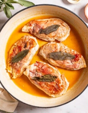 An overhead photo of chicken saltimbocca in a skillet. This is surrounded by sage, a bowl of salt, and a kitchen towel.