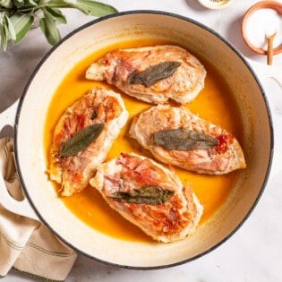 An overhead photo of chicken saltimbocca in a skillet. This is surrounded by sage, a bowl of salt, and a kitchen towel.