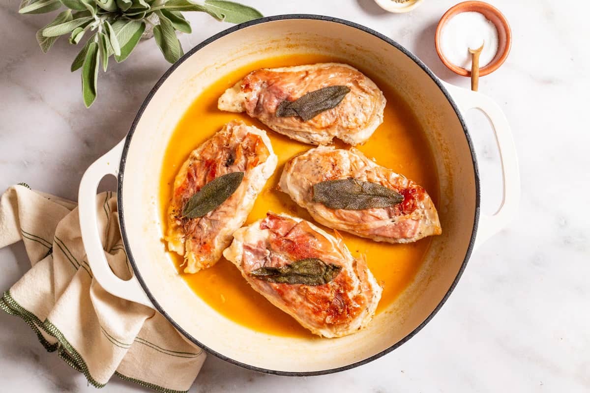 An overhead photo of chicken saltimbocca in a skillet. This is surrounded by sage, a bowl of salt, and a kitchen towel.