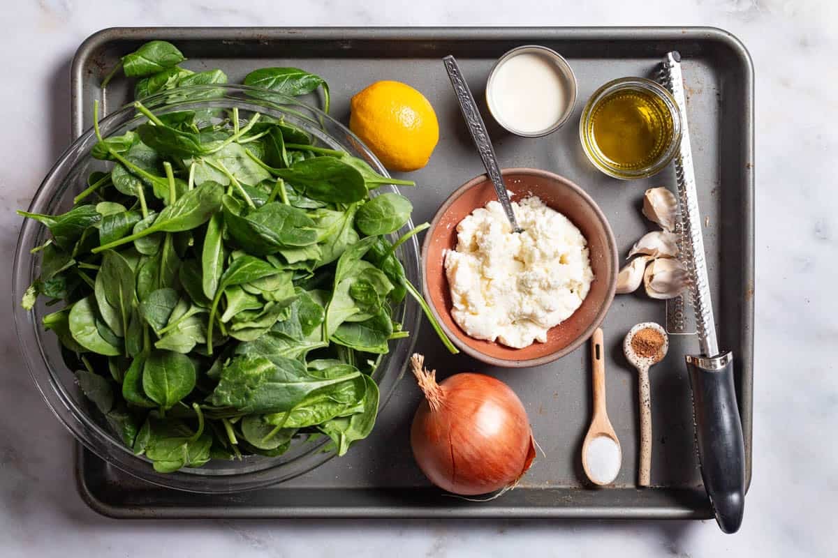 Ingredients for creamed spinach including fresh baby spinach, yellow onion, lemon, ricotta, milk, olive oil, garlic, salt and nutmeg.