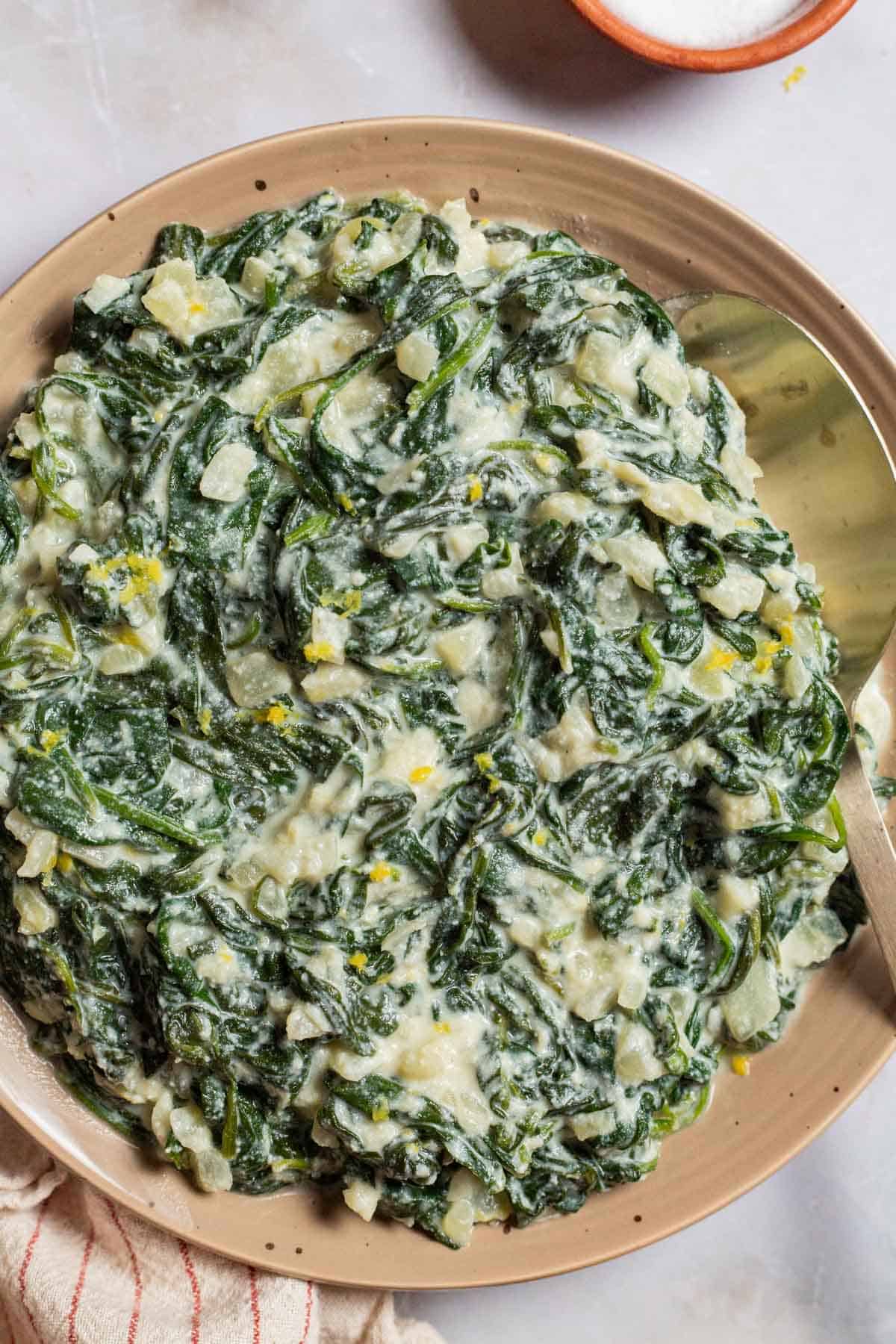 An overhead photo of creamed spinach on a plate with a spoon. Next to this is a small bowl of salt and a towel.