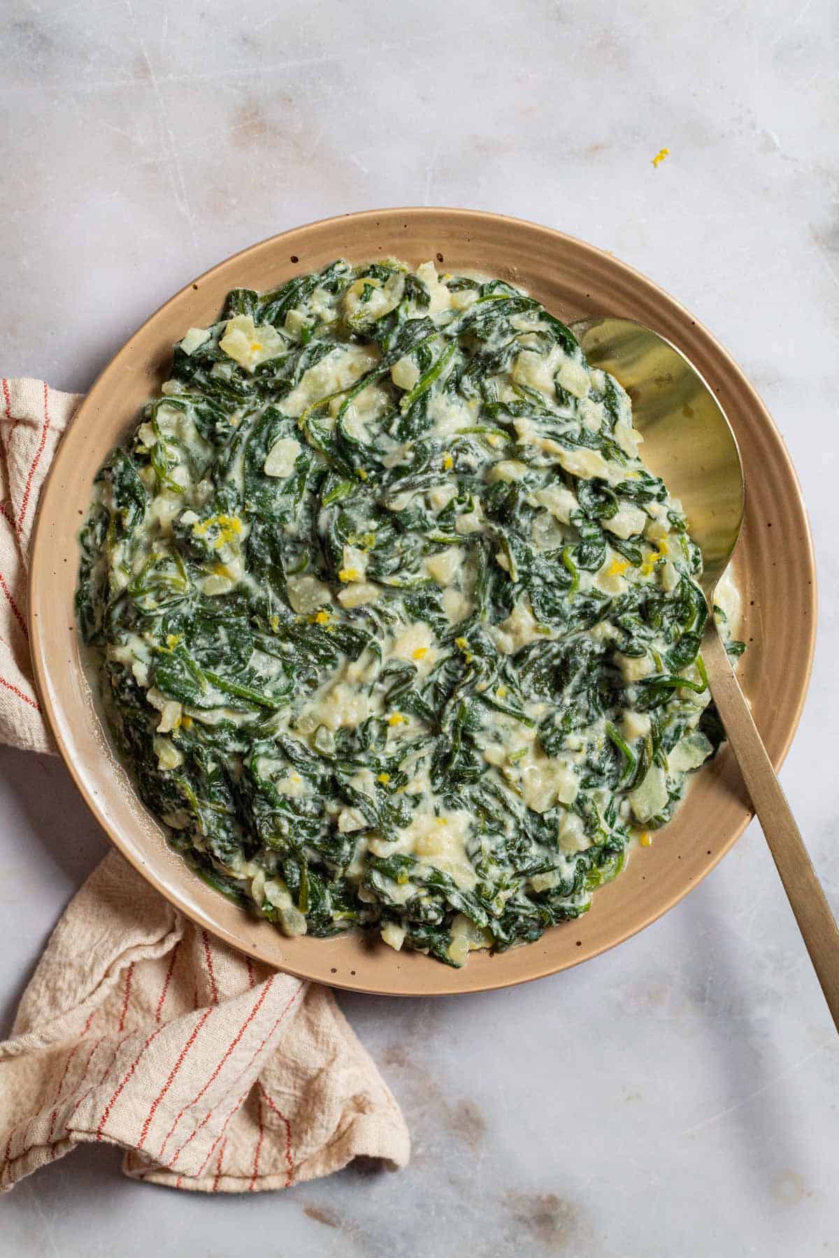 An overhead photo of creamed spinach on a plate with a spoon next to a towel.