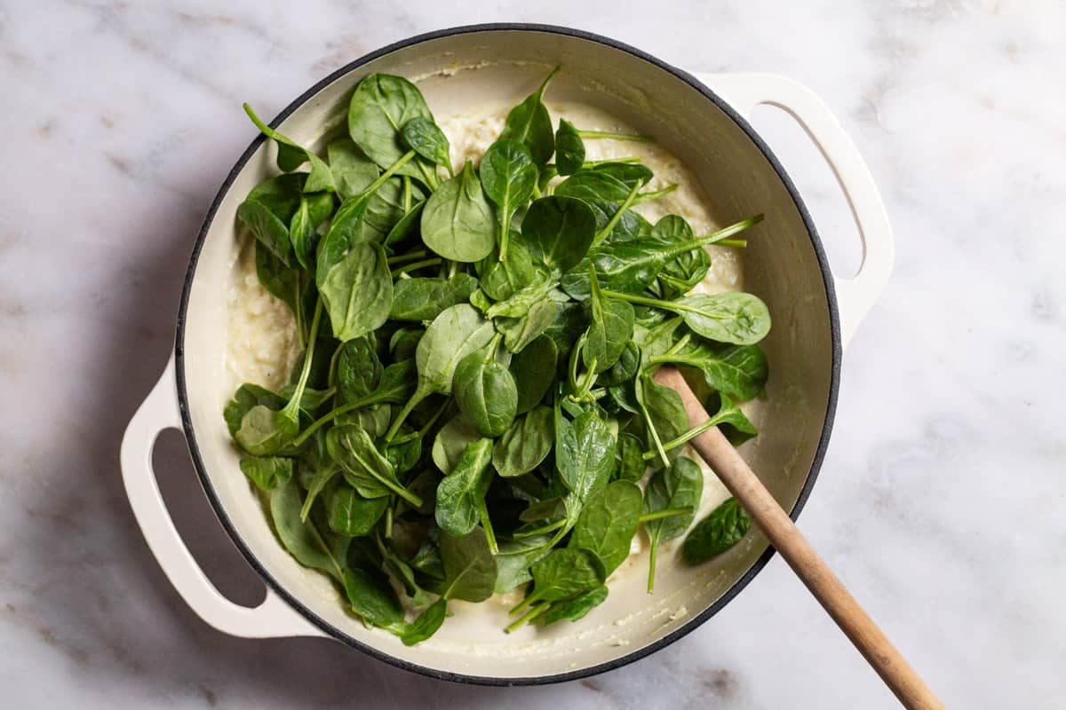Fresh spinach being added to the sauce in a skillet with a wooden spoon.