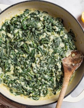 An overhead photo of creamed spinach in a skillet with a wooden spoon. Next to this is a towel, 2 lemon halves, and a small bowl of salt with a spoon.