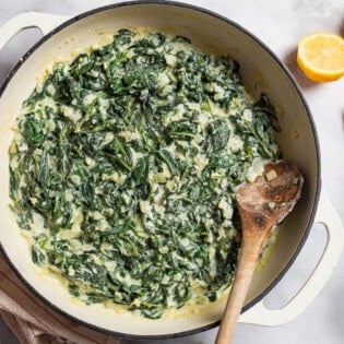 An overhead photo of creamed spinach in a skillet with a wooden spoon. Next to this is a towel, 2 lemon halves, and a small bowl of salt with a spoon.