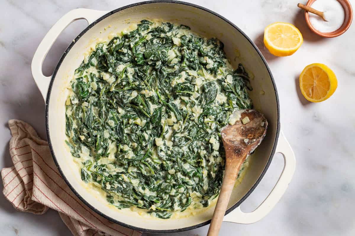 An overhead photo of creamed spinach in a skillet with a wooden spoon. Next to this is a towel, 2 lemon halves, and a small bowl of salt with a spoon.