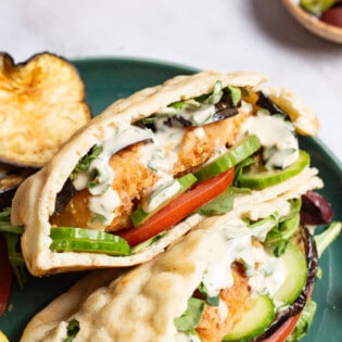 A close up of two fried fish sandwiches on a plate with sides of roasted eggplant slices, tomato, and a lemon wedge.