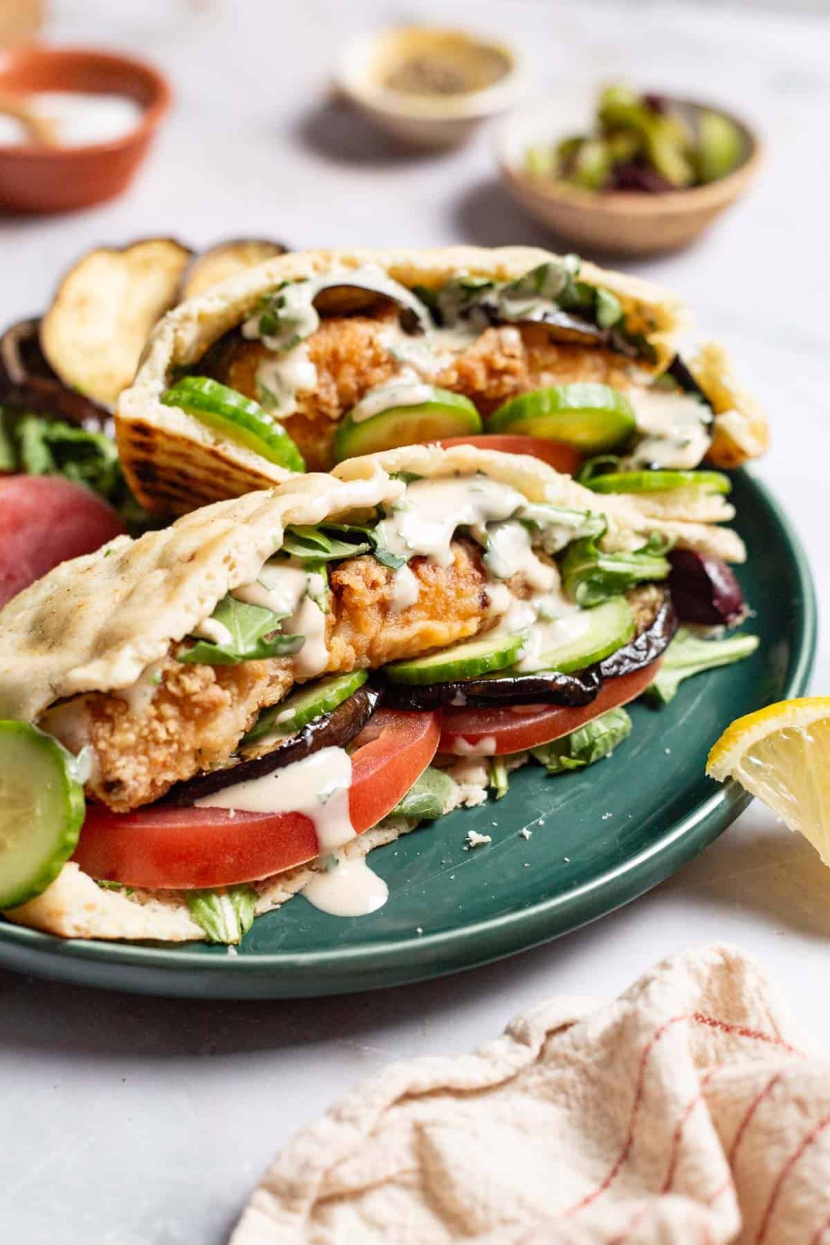 Two fried fish sandwiches on a plate with sides of roasted eggplant slices, tomato, persian cucumber slices and a lemon wedge. In the background are small bowls of salt, pepper and olives.