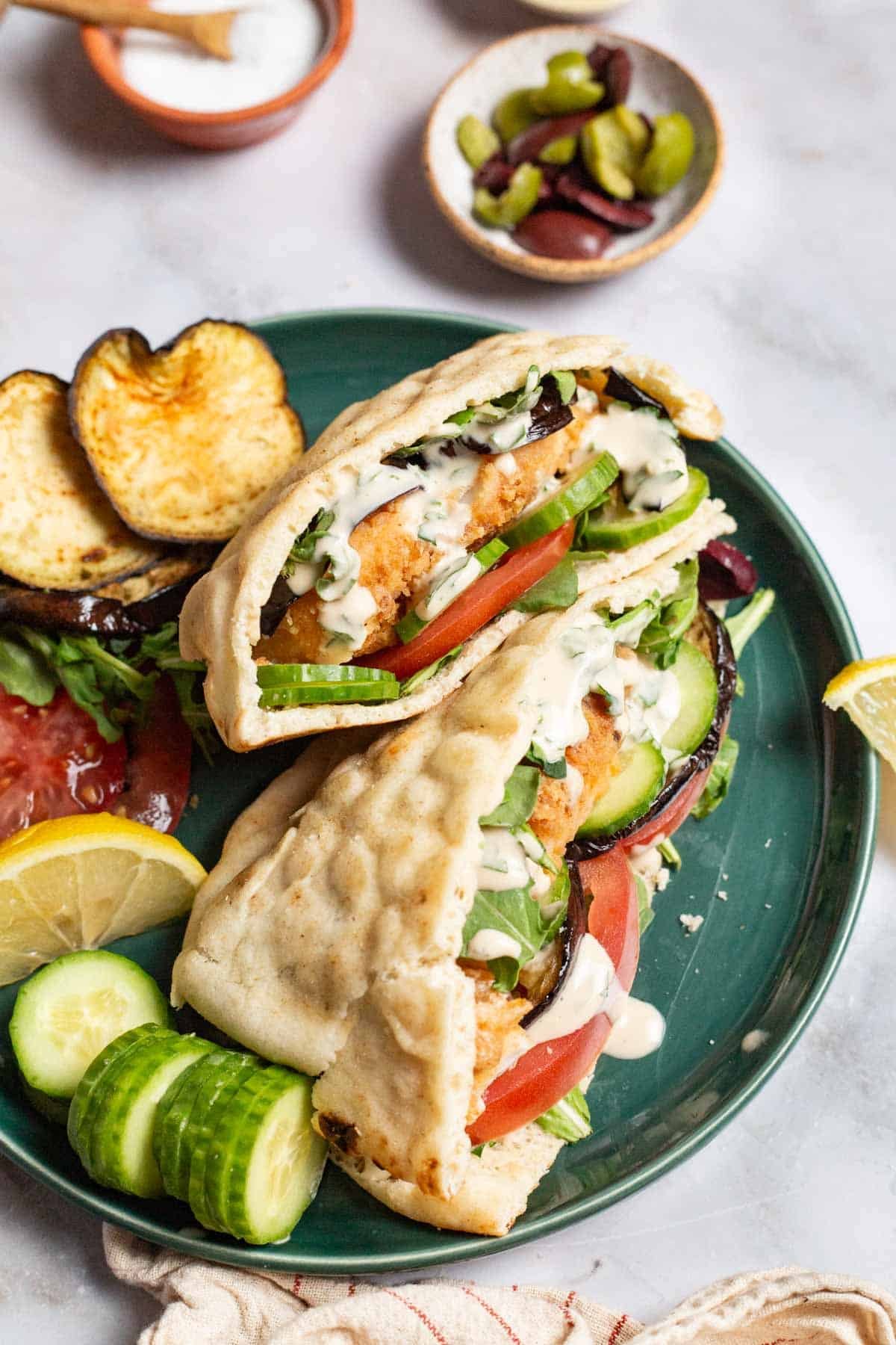 Two fried fish sandwiches on a plate with sides of roasted eggplant slices, tomato, persian cucumber slices and a lemon wedge. In the background is a small bowl of olives, and a small bowl of salt.