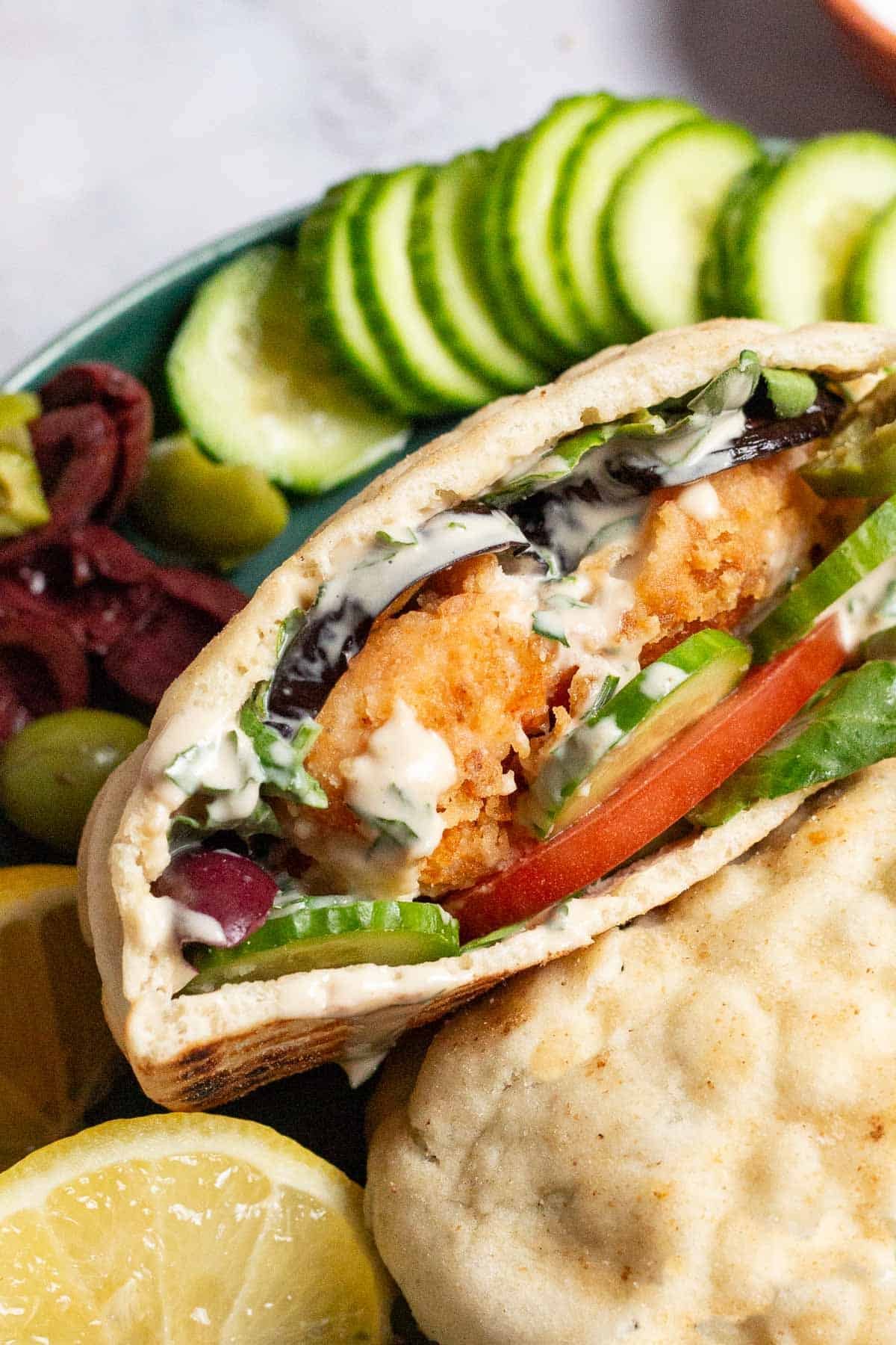 A close up of a fried fish sandwich on a plate surrounded by slices persian cucumbers, olives and a lemon half.