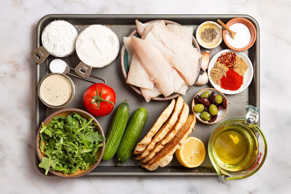 ingredients for fried fish sandwiches including white fish fillets, coriander, cumin, paprika, red pepper flakes, salt, pepper, garlic, lemon, olive oil, flour, corns starch, baking soda, persian cucumbers, tahini sauce, tomato, baby arugula, pita bread and olives.