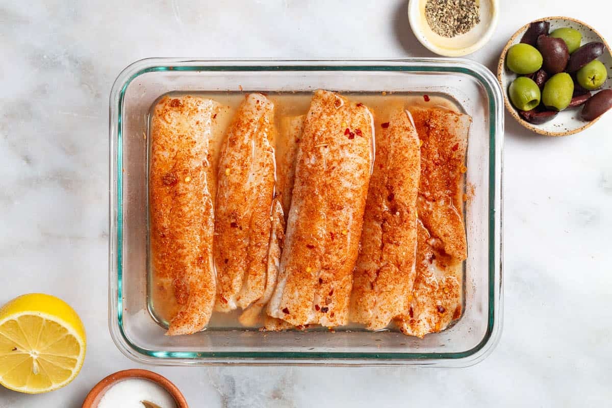 An overhead photo of seasoned, uncooked fish fillets in a baking dish surrounded by bowls of pepper, olives and salt, as well as a lemon halve.