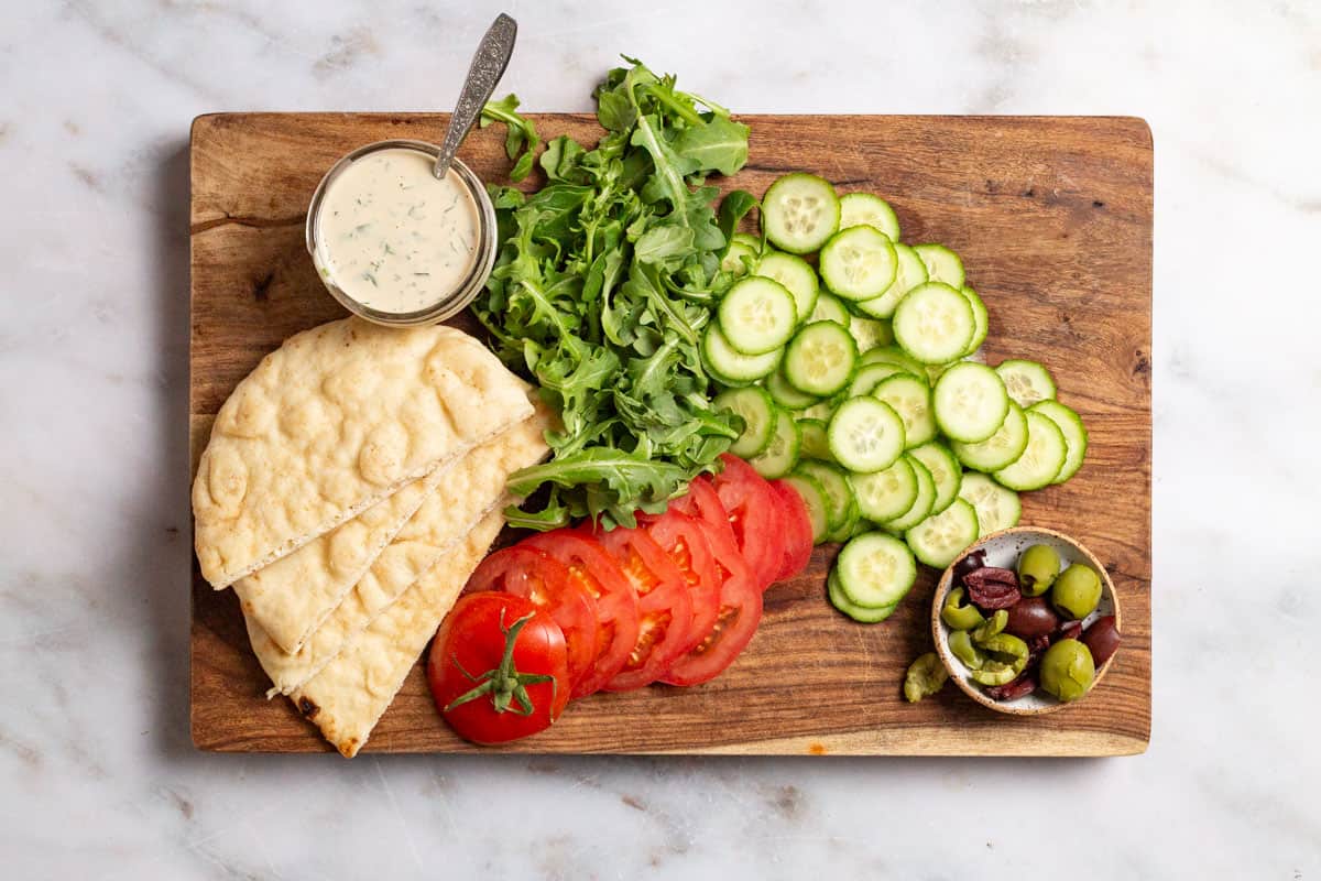 Fried fish fixings, including pita bread, tahini, arugula, slices persian cucumbers, sliced tomato and a bowl of olives on a wooden cutting board.