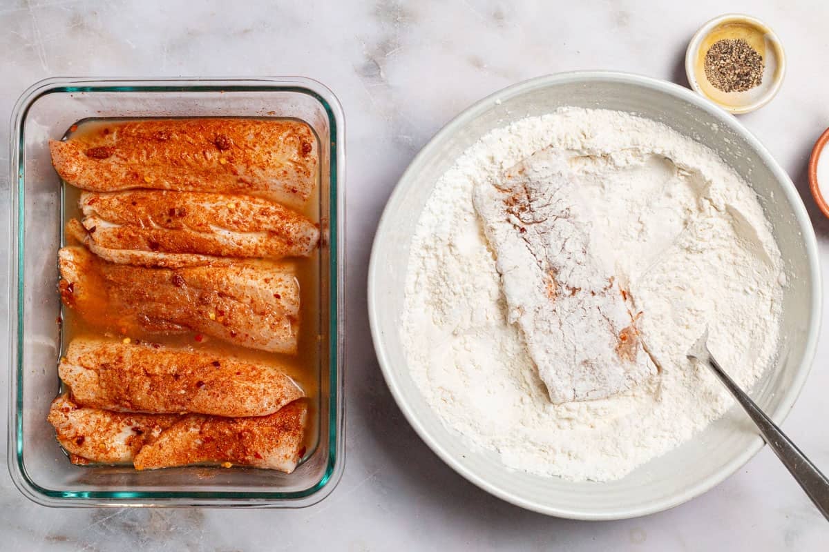 An overhead photo of seasoned, uncooked white fish fillets in a baking dish. Next to this is a fillet being dredged the flour mixture in a bowl with a fork, and a small bowl of pepper.