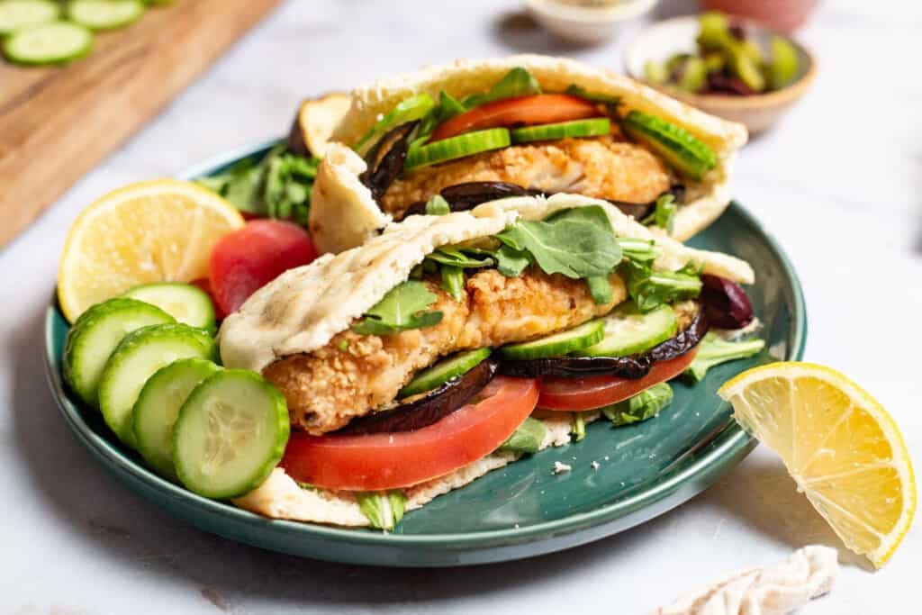 A close up of two fried fish sandwiches on a plate with sides of roasted eggplant slices tomato, persian cucumber slices and a lemon wedge. In the background is a small bowl of olives.