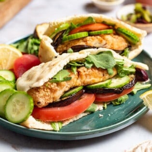 A close up of two fried fish sandwiches on a plate with sides of roasted eggplant slices tomato, persian cucumber slices and a lemon wedge. In the background is a small bowl of olives.