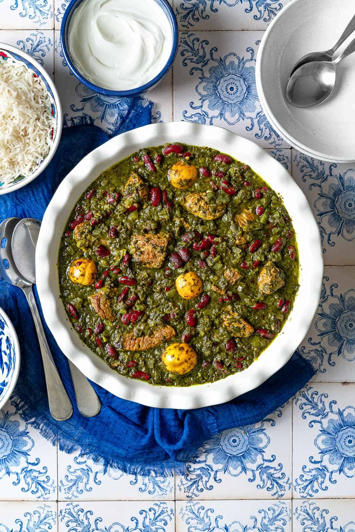 An overhead photo of ghormeh sabzi in a serving bowl. Next to this are serving utensils, bowls of rice and Greek yogurt, and empty bowls with 2 spoons.