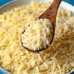Cooked orzo pasta on a wooden spoon being held about a bowl of orzo.