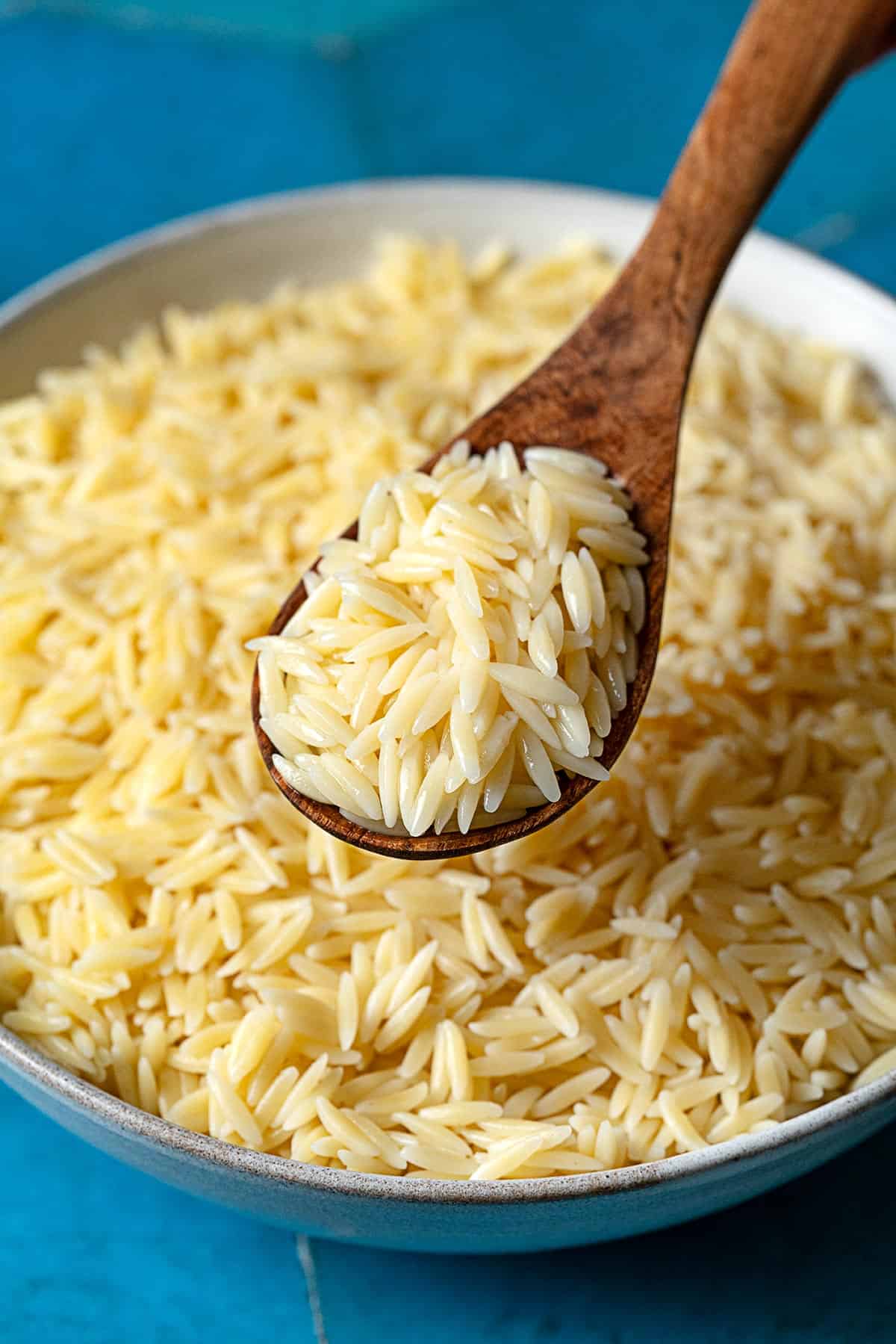 Cooked orzo pasta on a wooden spoon being held about a bowl of orzo.