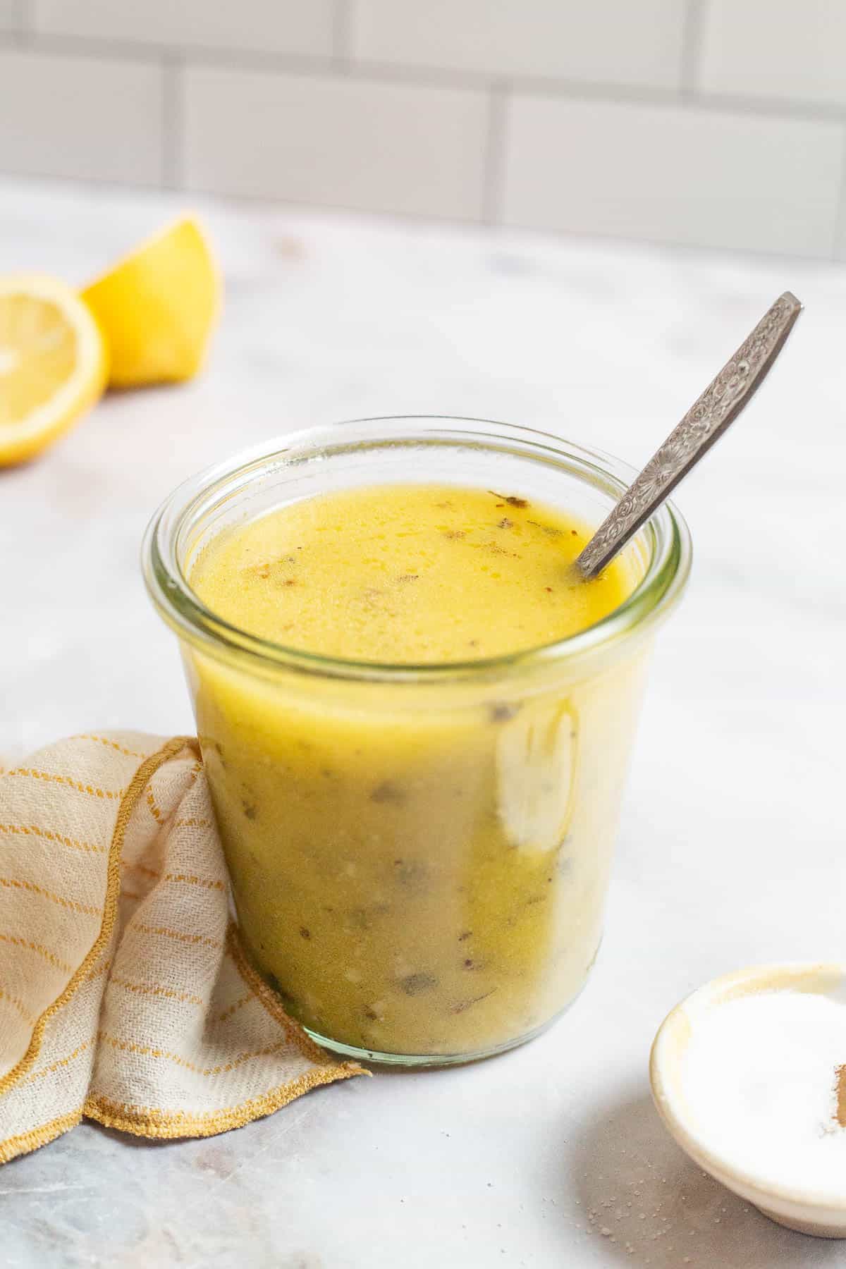 a jar of lemon vinaigrette with a spoon next to lemon halves, a small bowl of salt, and a cloth towel.