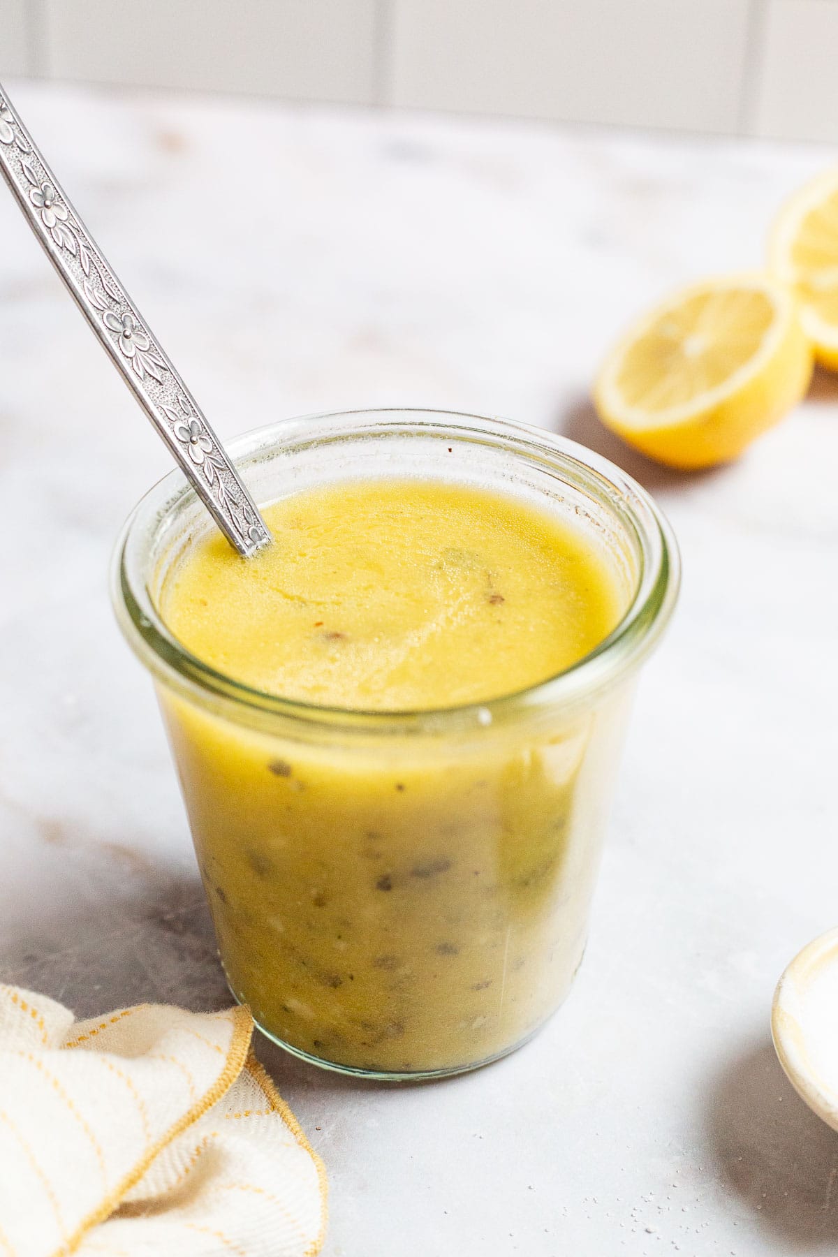 a jar of lemon vinaigrette with a spoon next to lemon halves and a cloth towel.