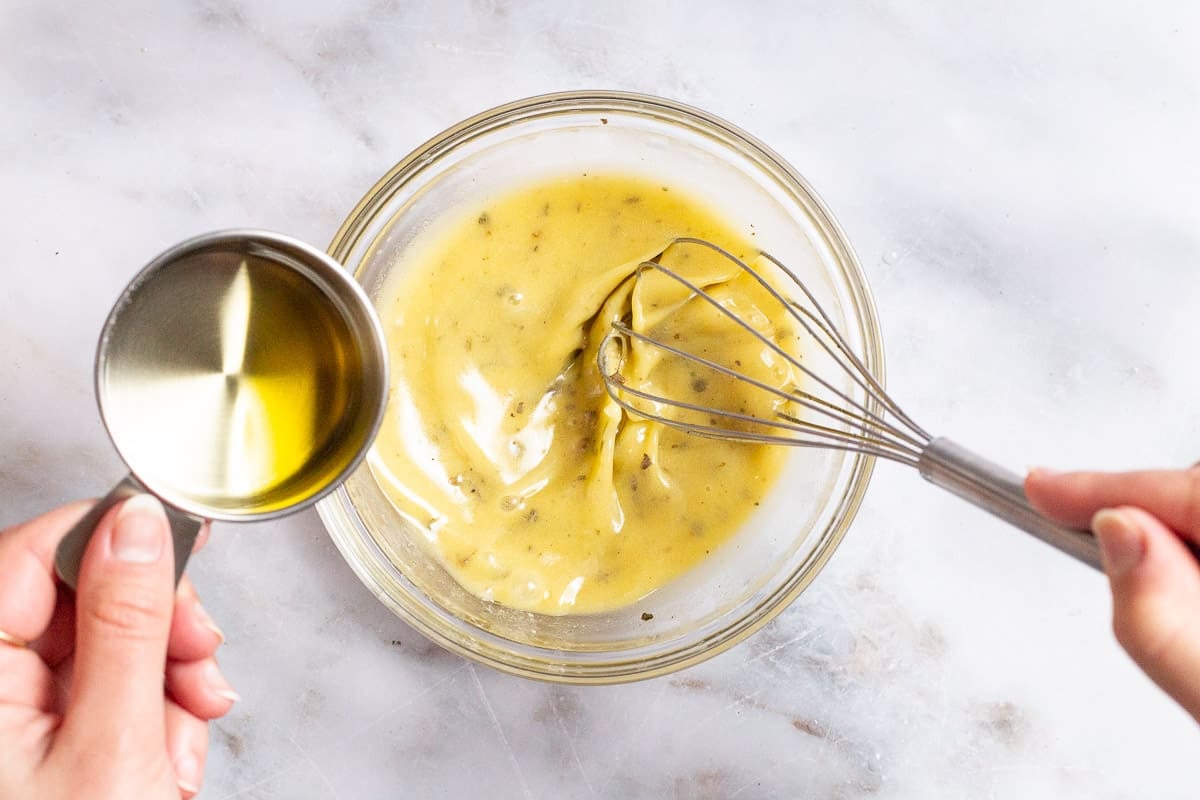 a cup of olive oil slowly being added to the bowl of the lemon vinaigrette as it's being whisked.