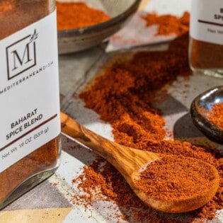 Baharat spice blend on a spoon and in a small bowl. Next to this are two jars of the spice from The Mediterranean Dish.