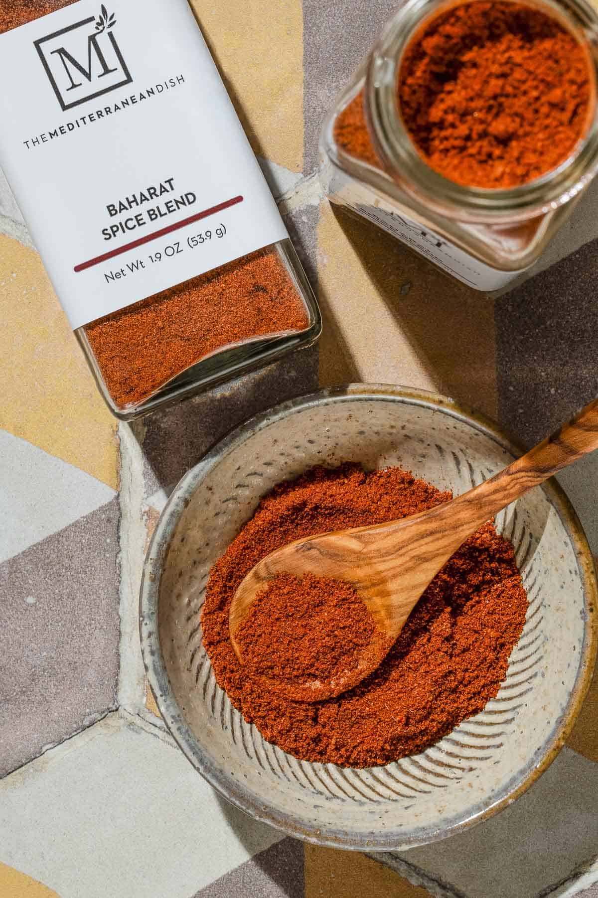 An overhead photo of baharat spice blend in a bowl with a small wooden spoon. Next to this are two jars of the spice from The Mediterranean Dish.