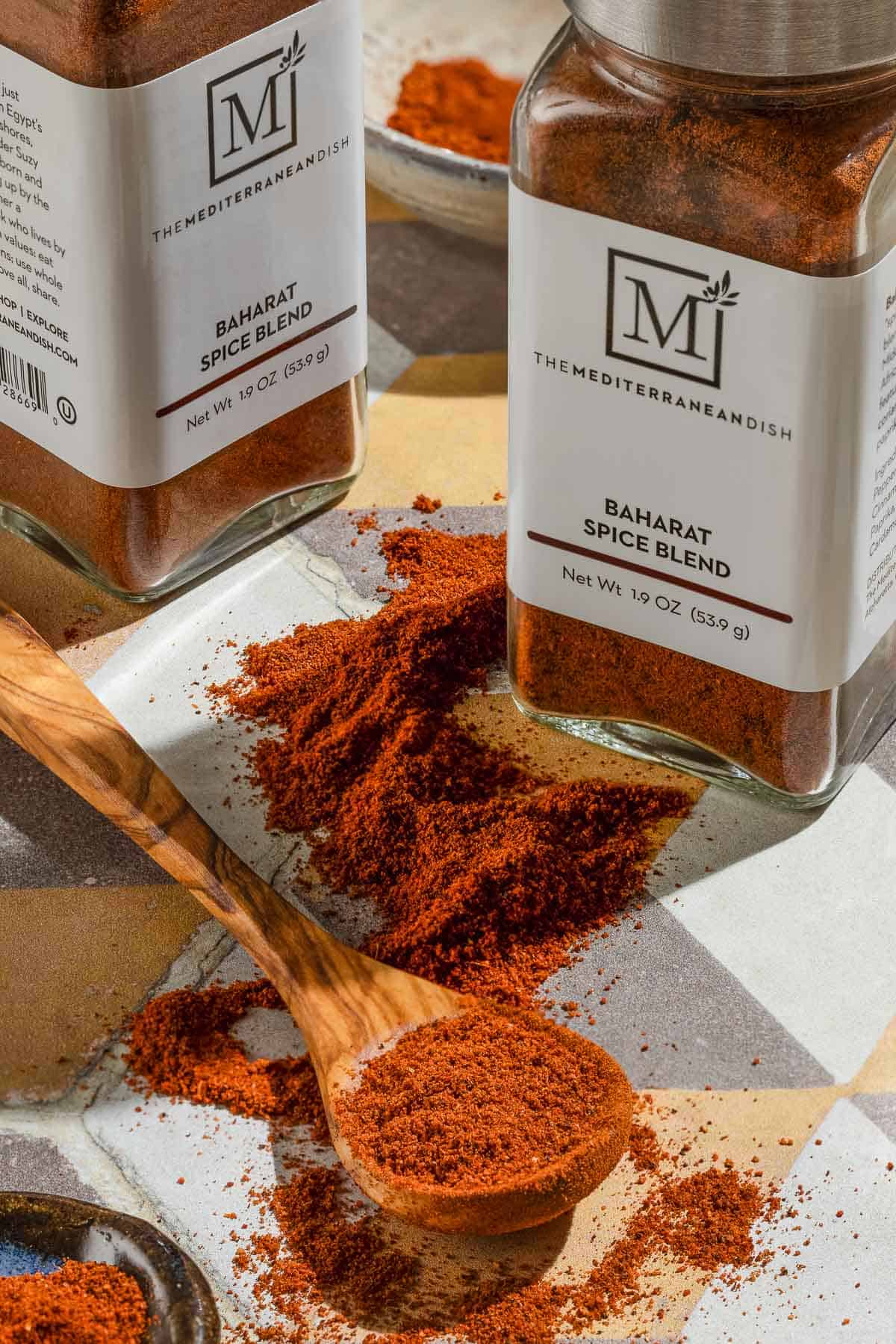 Baharat spice blend on a spoon and in a small bowl. Next to this are two jars of the spice from The Mediterranean Dish.