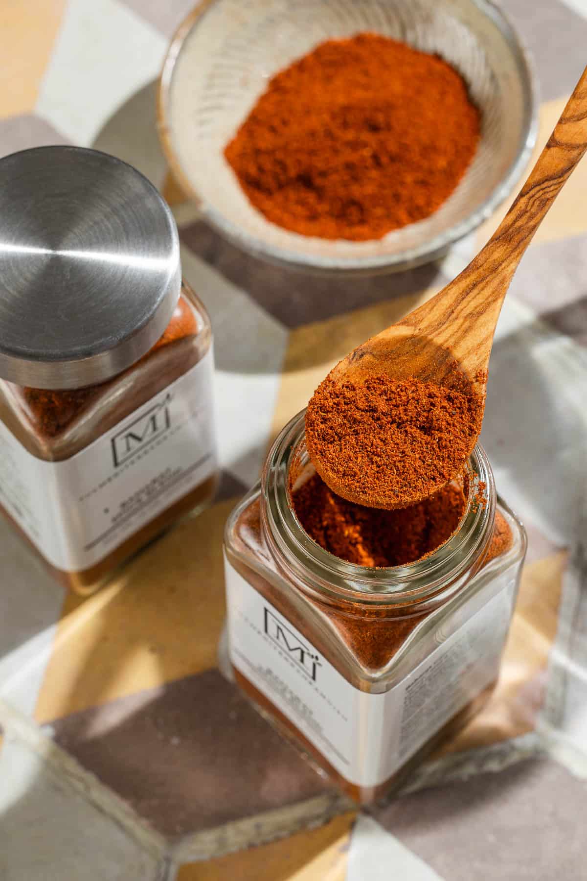 Baharat spice blend on a small wooden spoon being held over an open jar of the spice. Next to this is another jar and a small bowl of the spice.