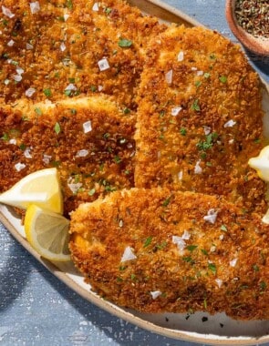 An overhead photo of 4 chicken cutlets on a platter with lemon wedges. Next to this is a glass of wine and small bowls of Italian seasoning and salt.