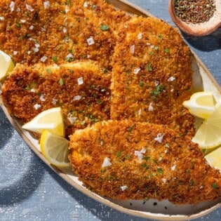 An overhead photo of 4 chicken cutlets on a platter with lemon wedges. Next to this is a glass of wine and small bowls of Italian seasoning and salt.