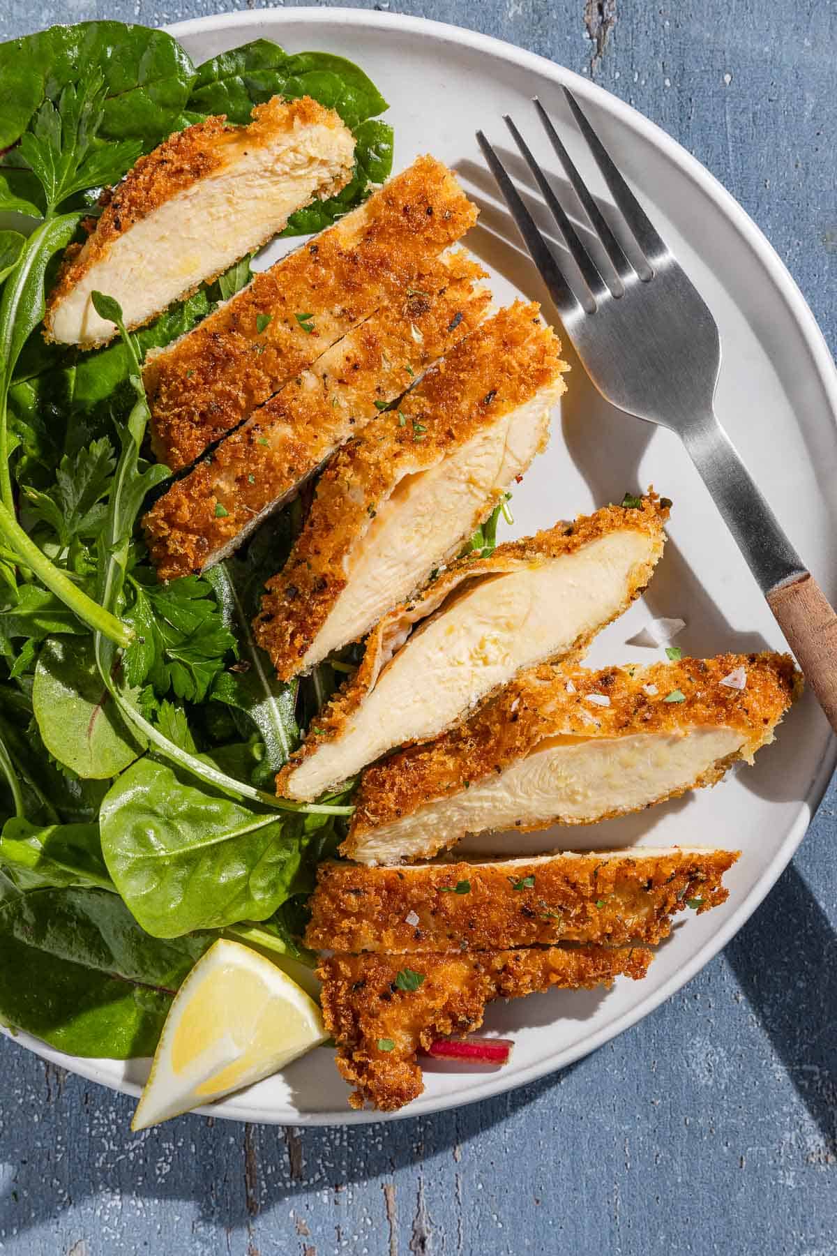 An overhead photo of a sliced chicken cutlet on a plate with a salad, a lemon wedge and a fork.