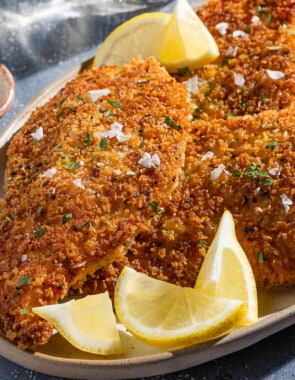 A close up of 4 chicken cutlets on a platter with lemon wedges next to small bowls of Italian seasoning and salt.