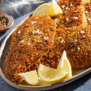 A close up of 4 chicken cutlets on a platter with lemon wedges next to small bowls of Italian seasoning and salt.