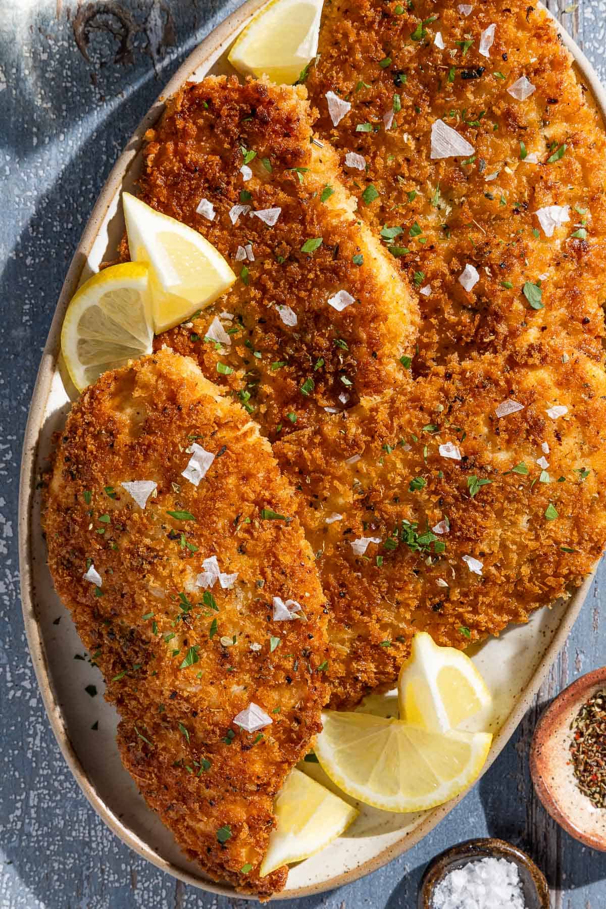 An overhead photo of 4 chicken cutlets on a platter with lemon wedges next to small bowls of Italian seasoning and salt.