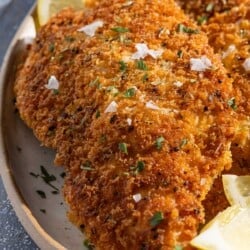 A close up of a chicken cutlet on a platter with lemon wedges.