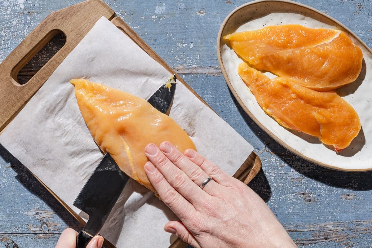 A chicken breast being cut into cutlets with a knife on a parchment lined cutting board. Next to this is a platter with 2 chicken cutlets.