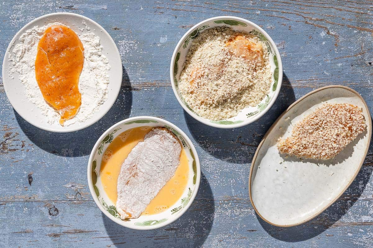 4 chicken cutlets going through the dredging process. One in a bowl of flour, one in a bowl of eggs, one in a bowl of bread crumbs, and one completed on a platter.