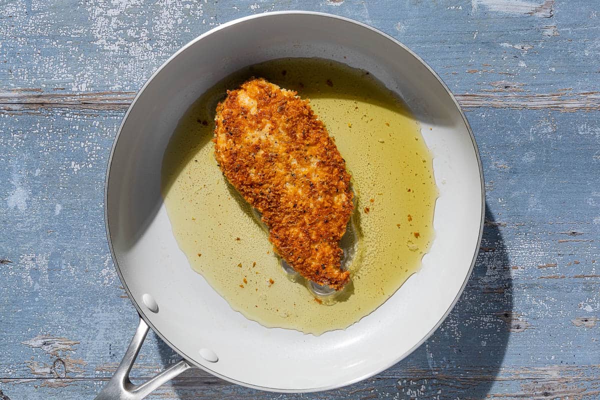 A breaded chicken cutlet being fried in olive oil in a skillet.