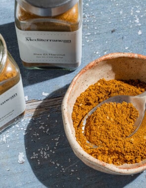 Ras el hanout in a bowl with a spoon. Next to this are two jars of ras el hanout from the mediterranean dish.