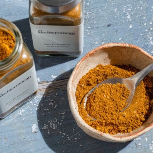 Ras el hanout in a bowl with a spoon. Next to this are two jars of ras el hanout from the mediterranean dish.