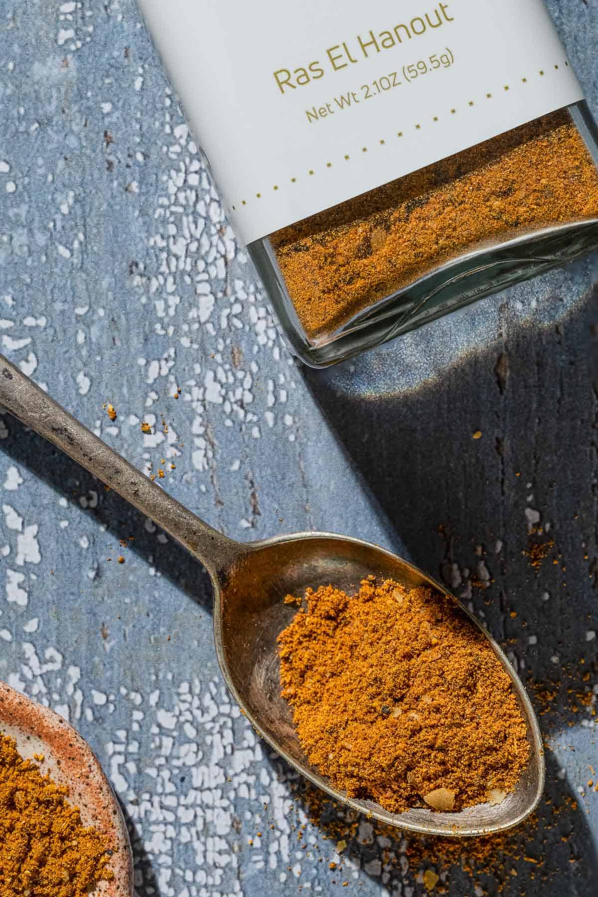 An overhead photo of ras el hanout on a spoon next to a jar of the spice from the mediterranean dish.